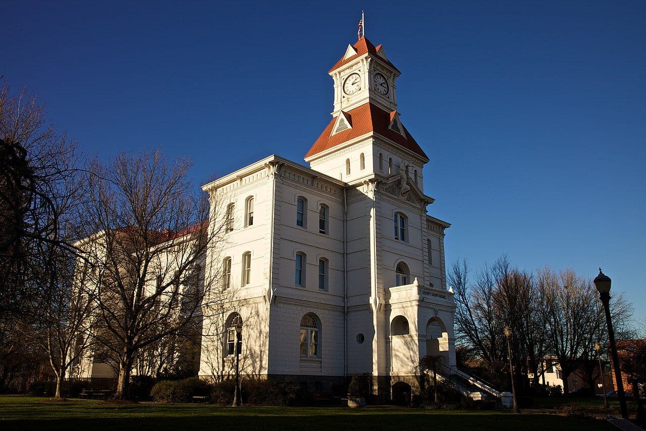 The white Benton County Courthouse and its clock tower in downtown Corvallis under a clear blue sky.